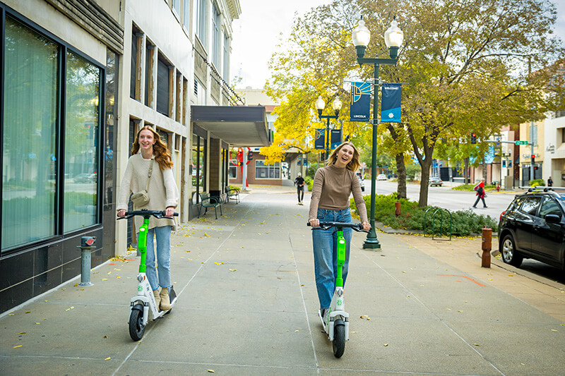 Two women ride scooters down the sidewalk.