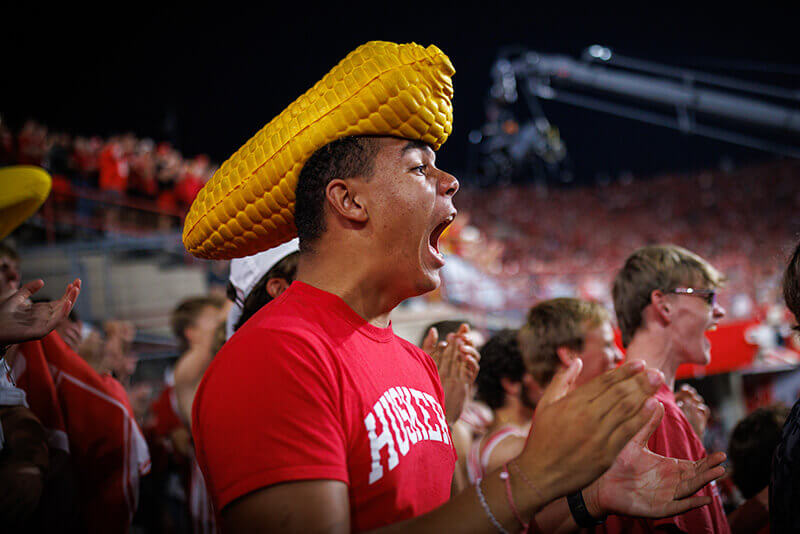 A diehard Husker fan wears a corn cob hat while yelling for his favorite team.